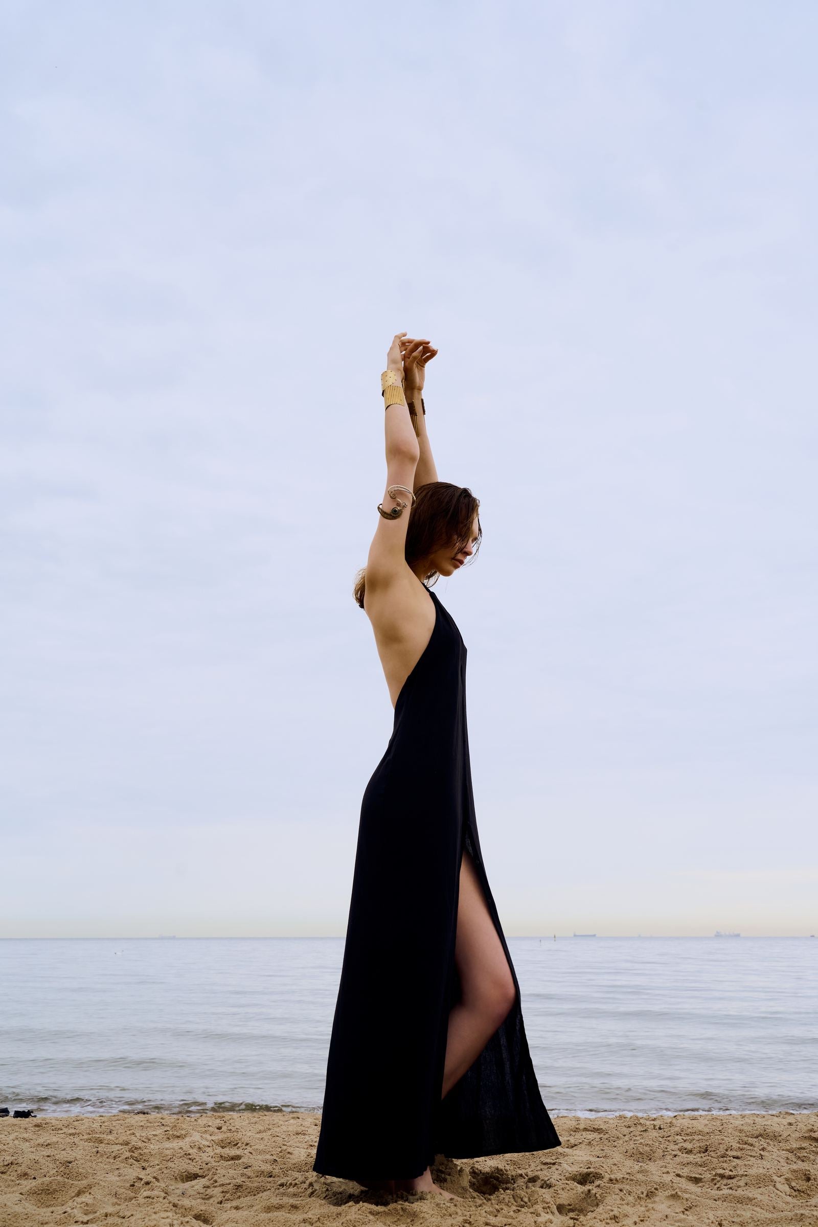 Woman in a black dress standing on a beach with arms raised against a clear sky.