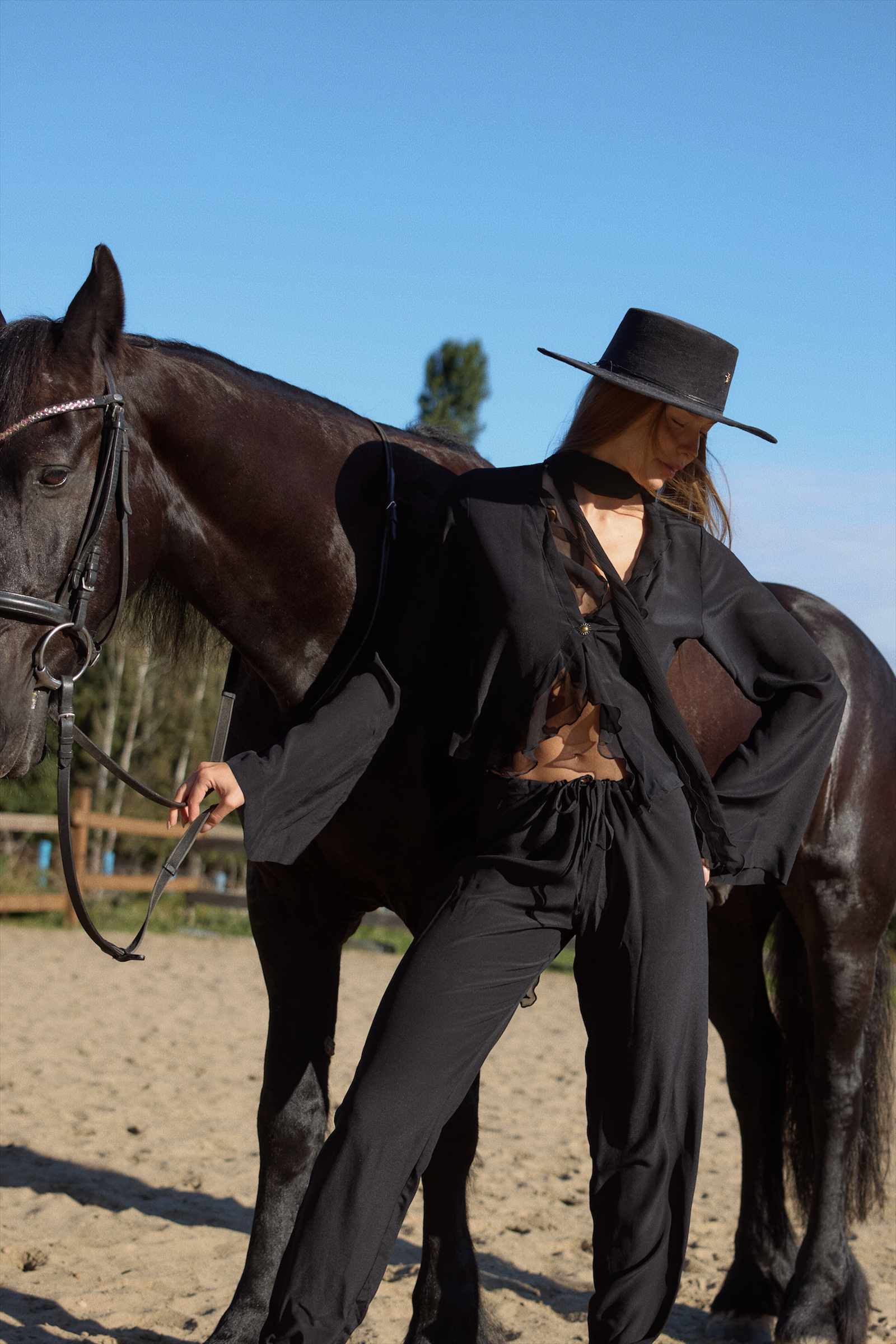 Person in black outfit and hat standing next to a horse on a sandy ground with trees in the background.