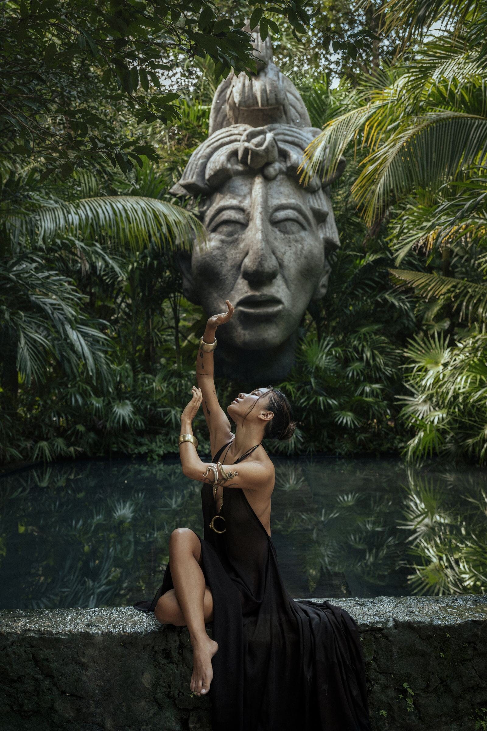 Woman in a brown dress sitting on a ledge with a large stone mask above her head, surrounded by lush greenery.