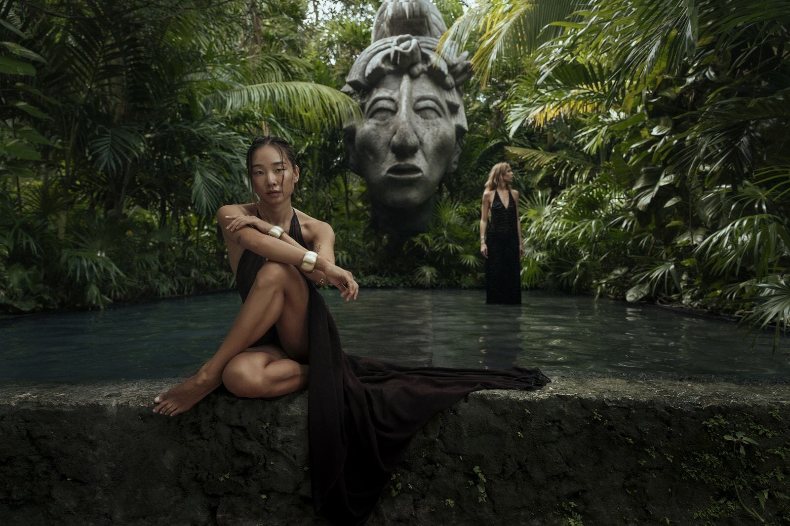 Two women in dark dresses sitting on a stone ledge with a large statue in the background amidst lush greenery.