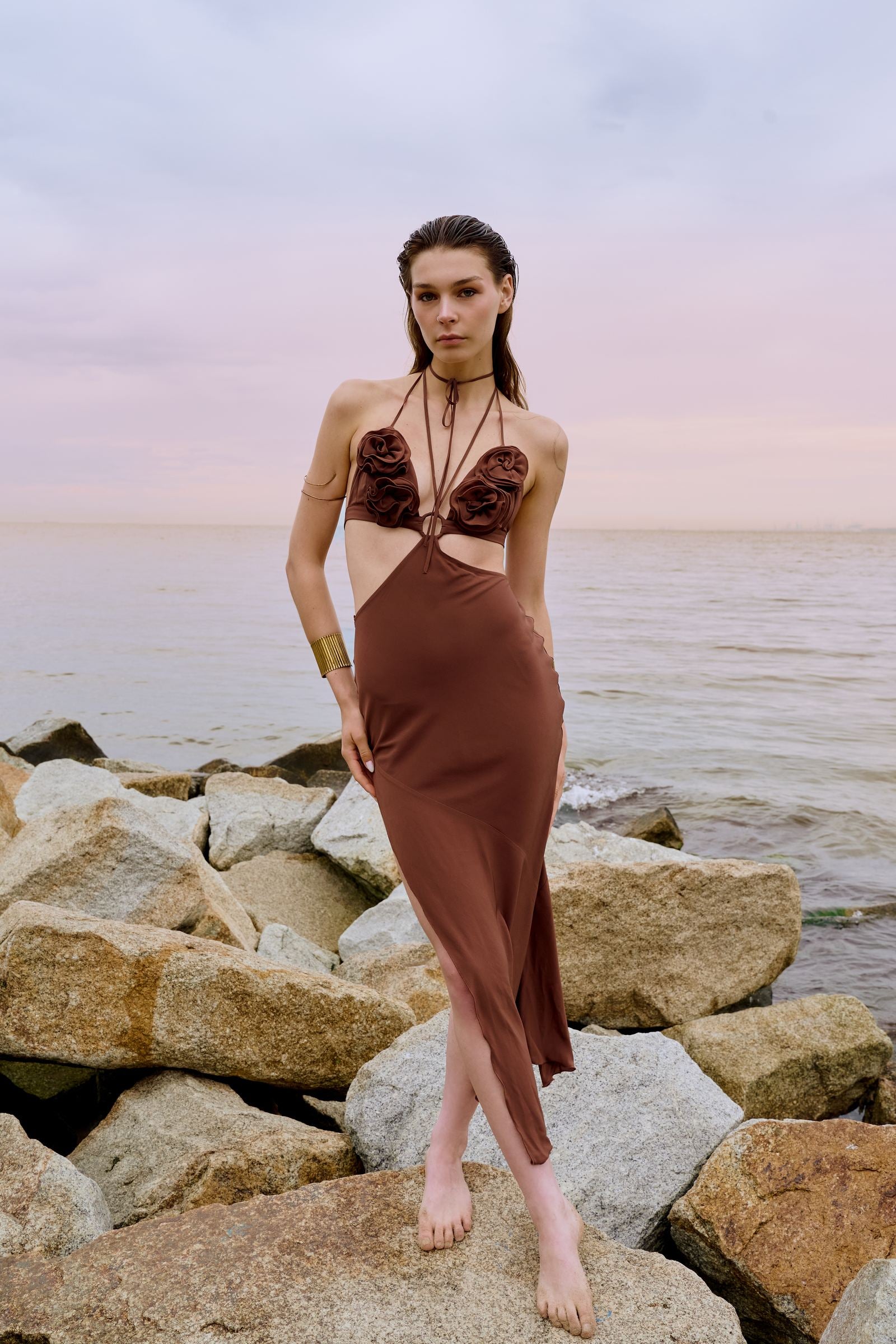 Woman in a brown dress standing on rocks by the water at sunset