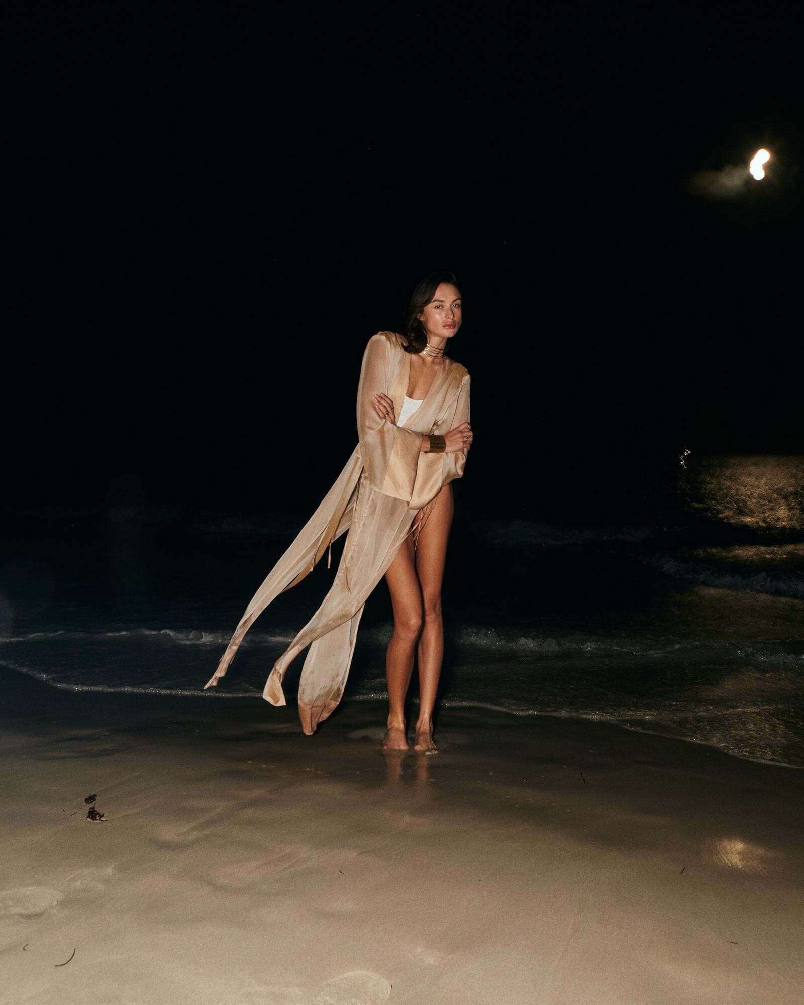 Woman in a flowing beige dress standing on a beach at night.