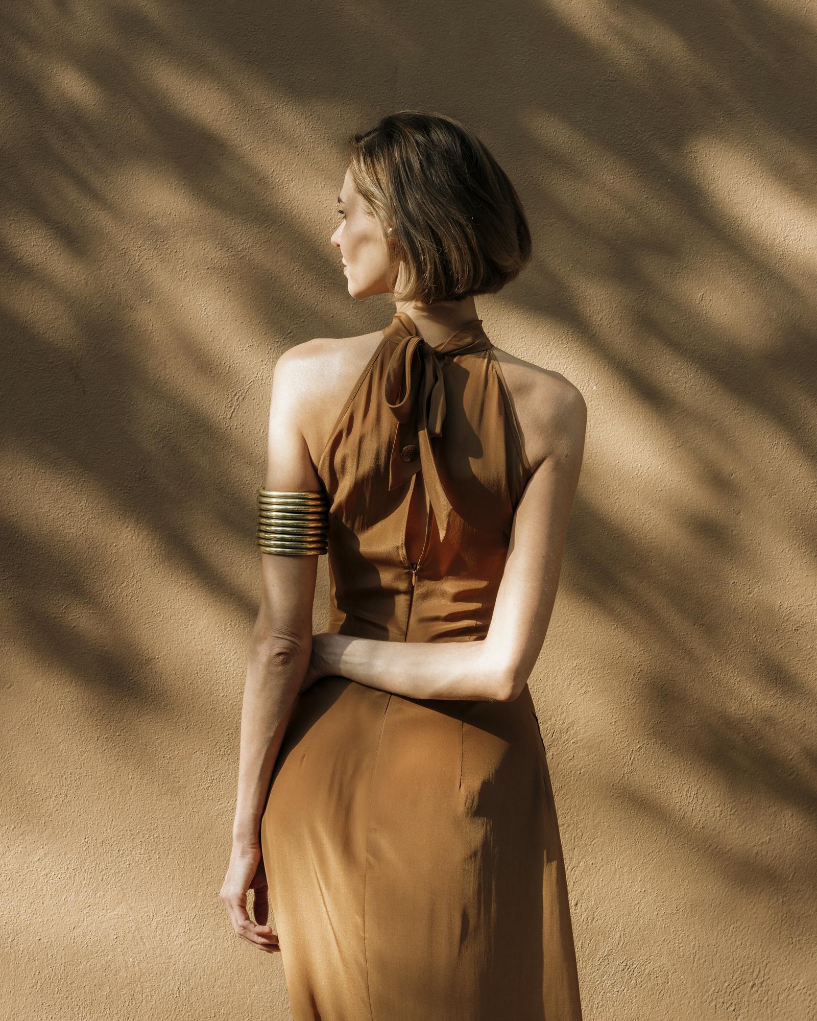 Woman in a golden dress standing against a textured beige wall with shadow patterns.