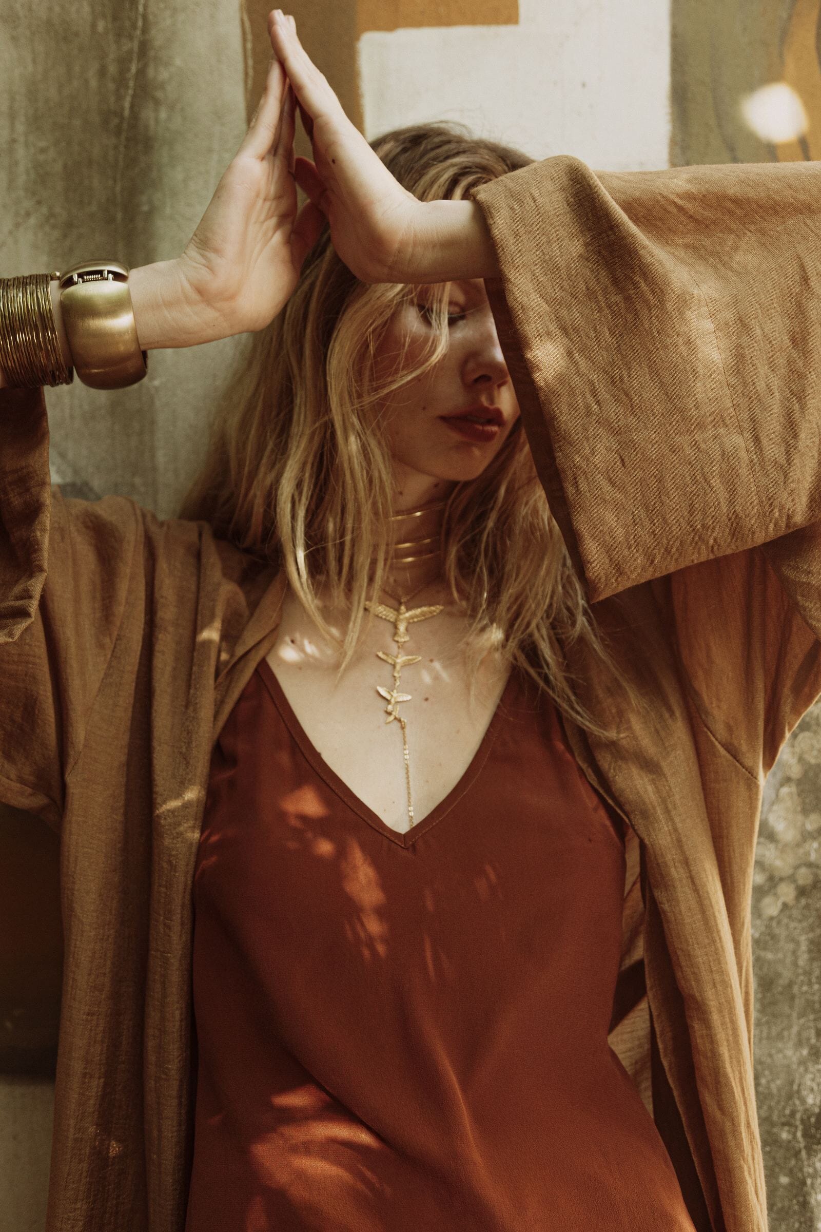 Woman in a gold dress with a beige cardigan holding her hands above her head, with a warm-toned filter.