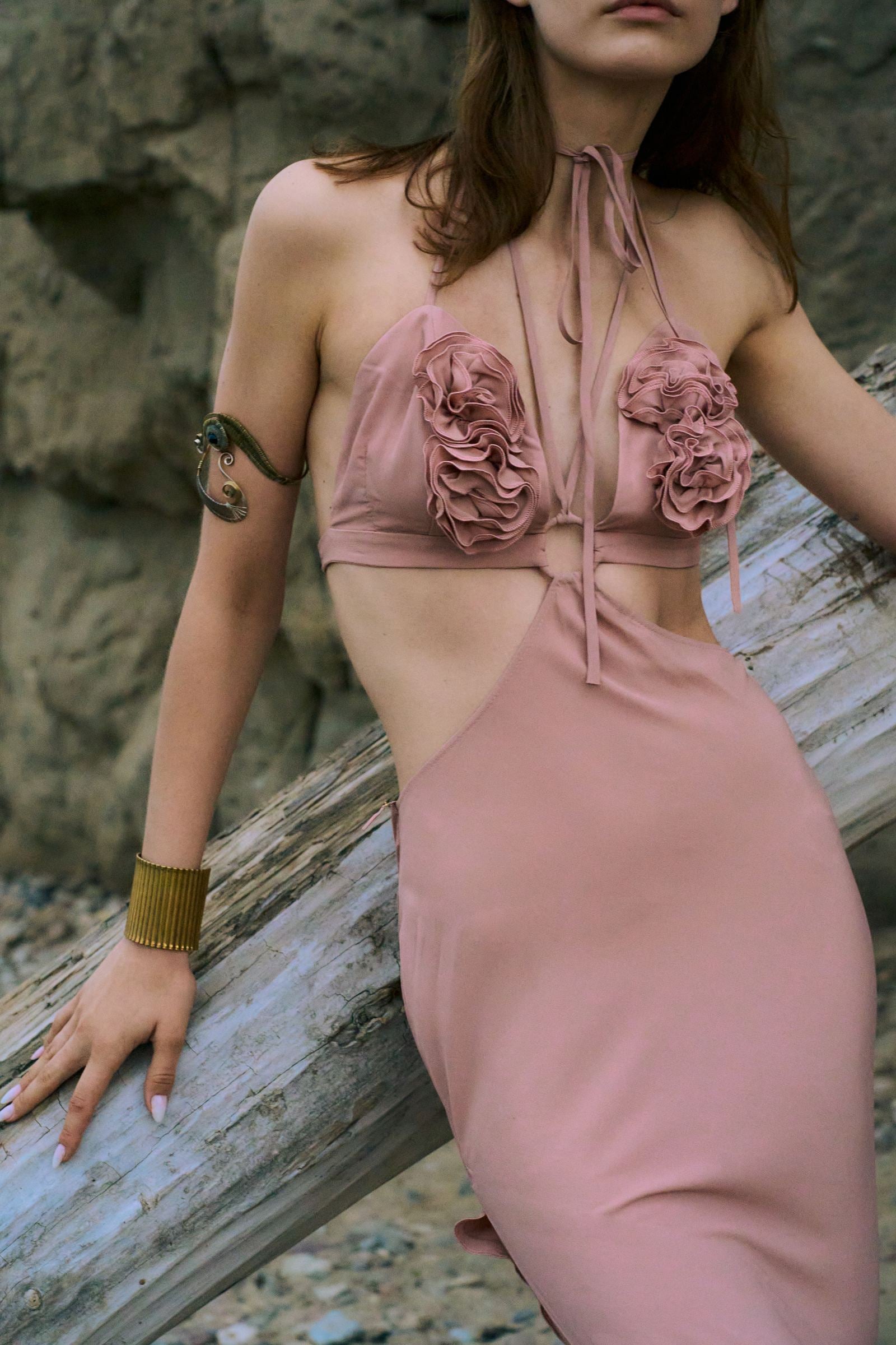 Woman wearing a pink floral dress leaning against a wooden log with a natural stone background.