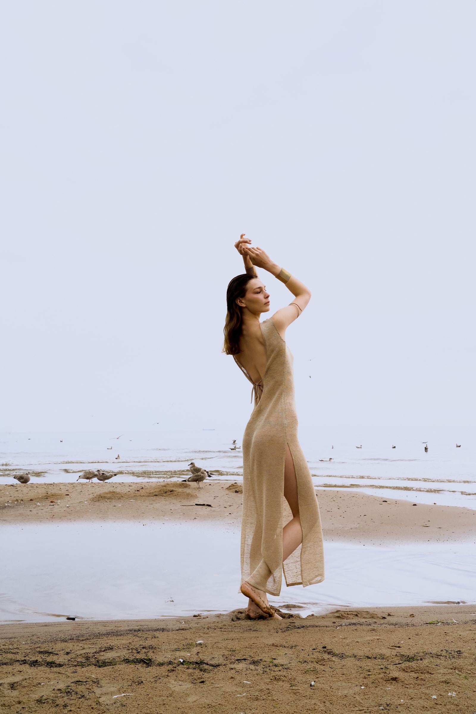 Woman in a long beige dress standing on a beach with a clear sky.