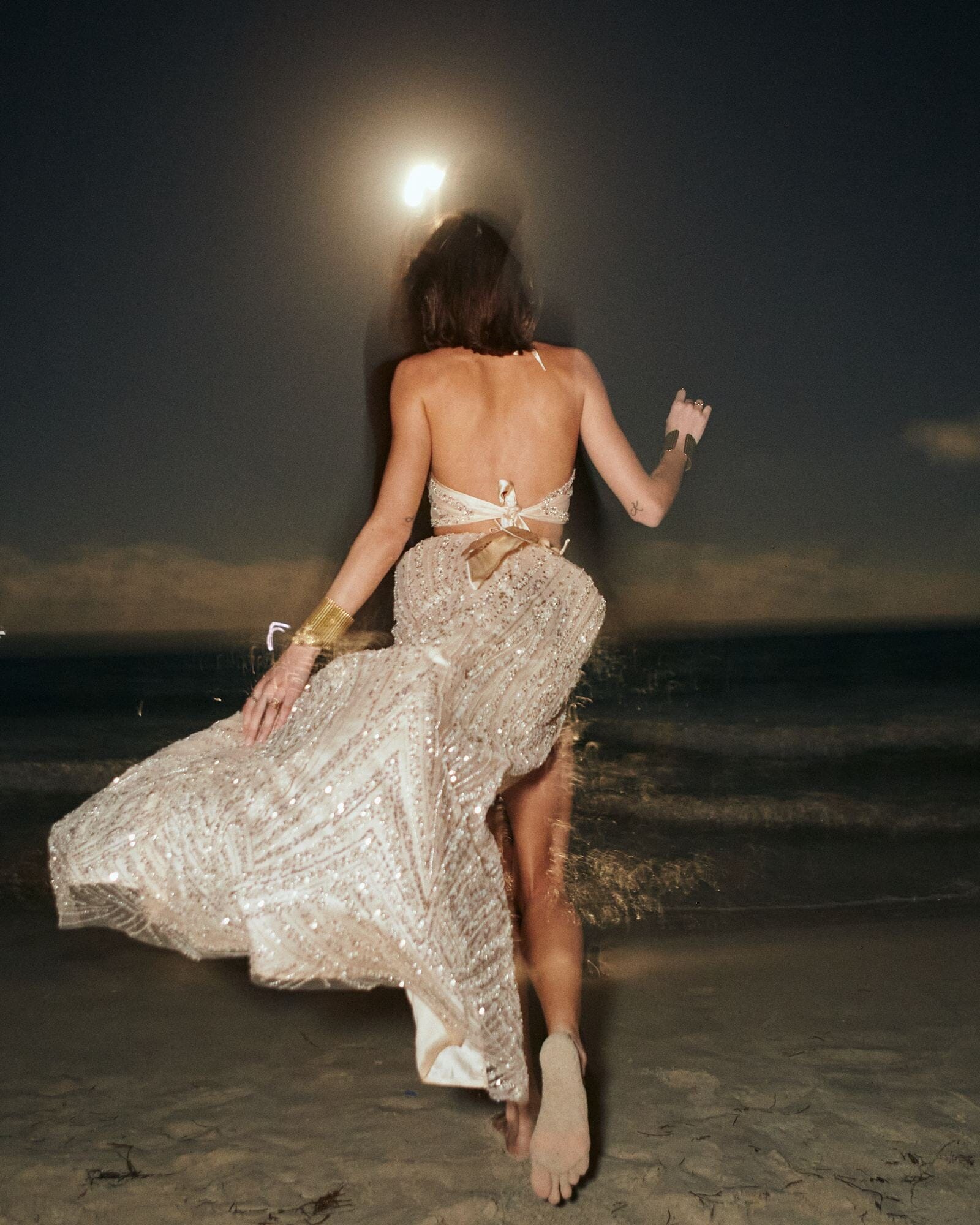 Woman in a sparkly dress running on a beach at night with a full moon.