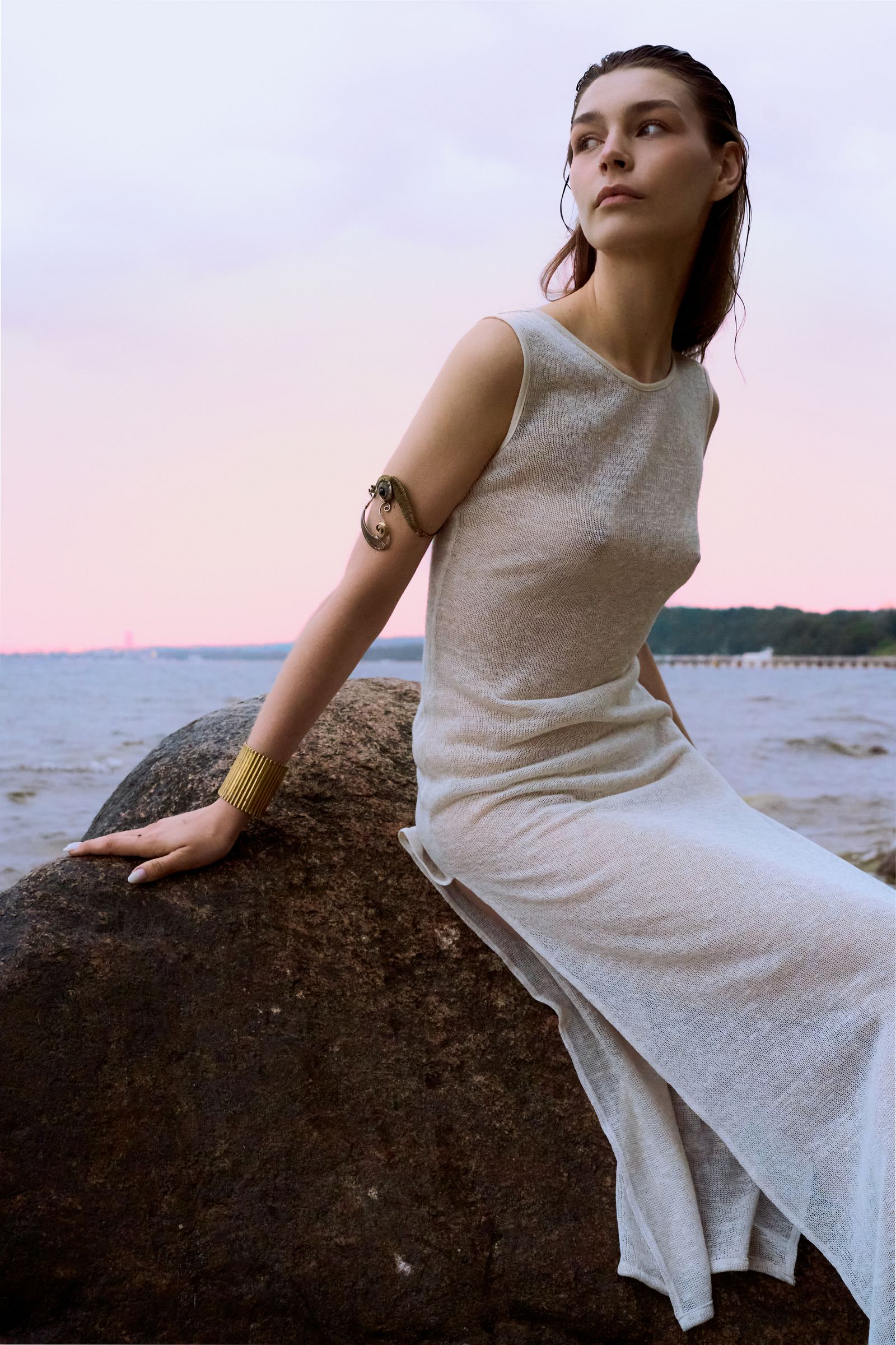 Woman in a white dress sitting on a rock by a body of water at sunset.