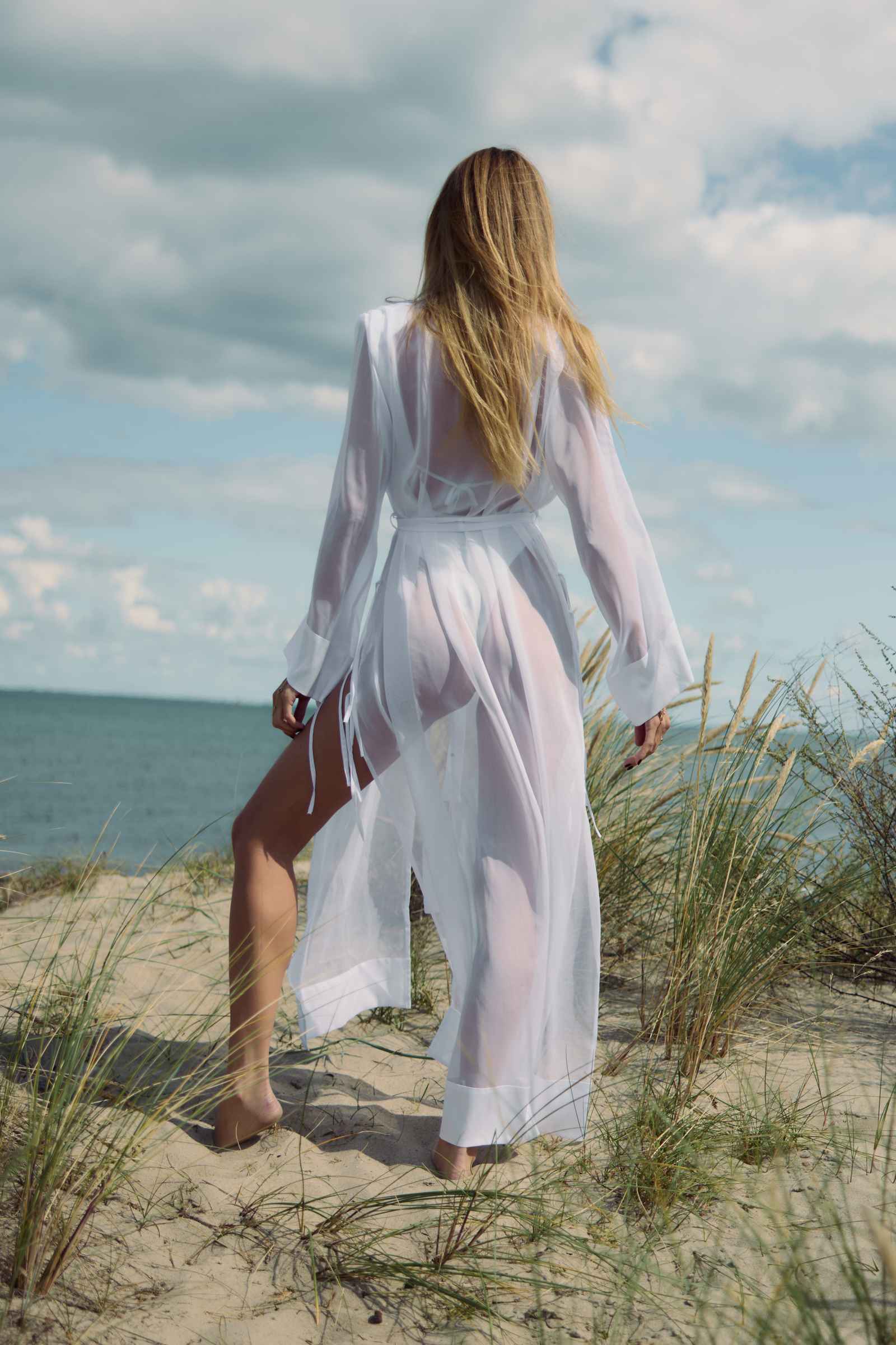 Woman in a white dress standing on a sandy beach with grass and ocean in the background
