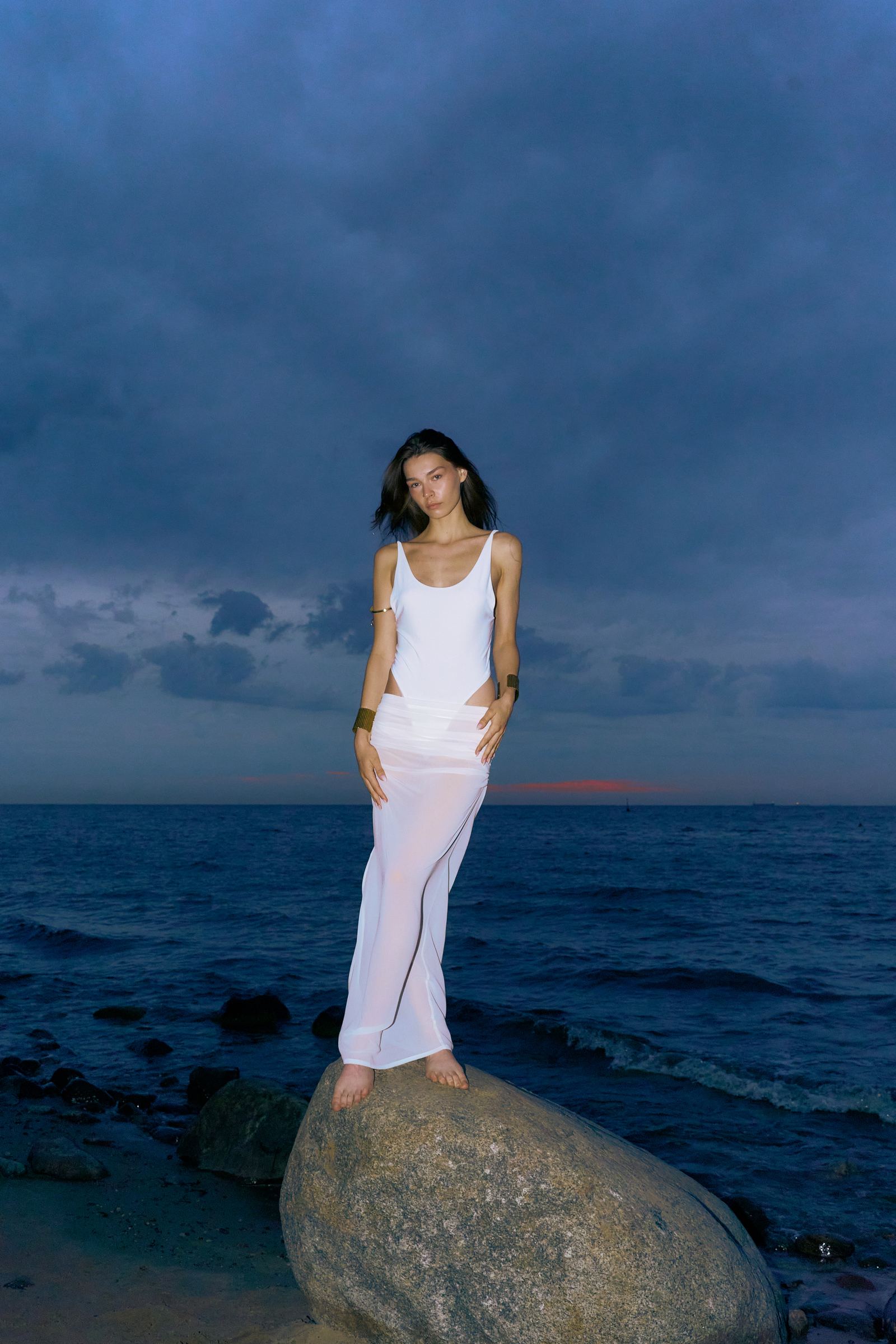 Woman in white outfit standing on a rock by the ocean under a dark sky.