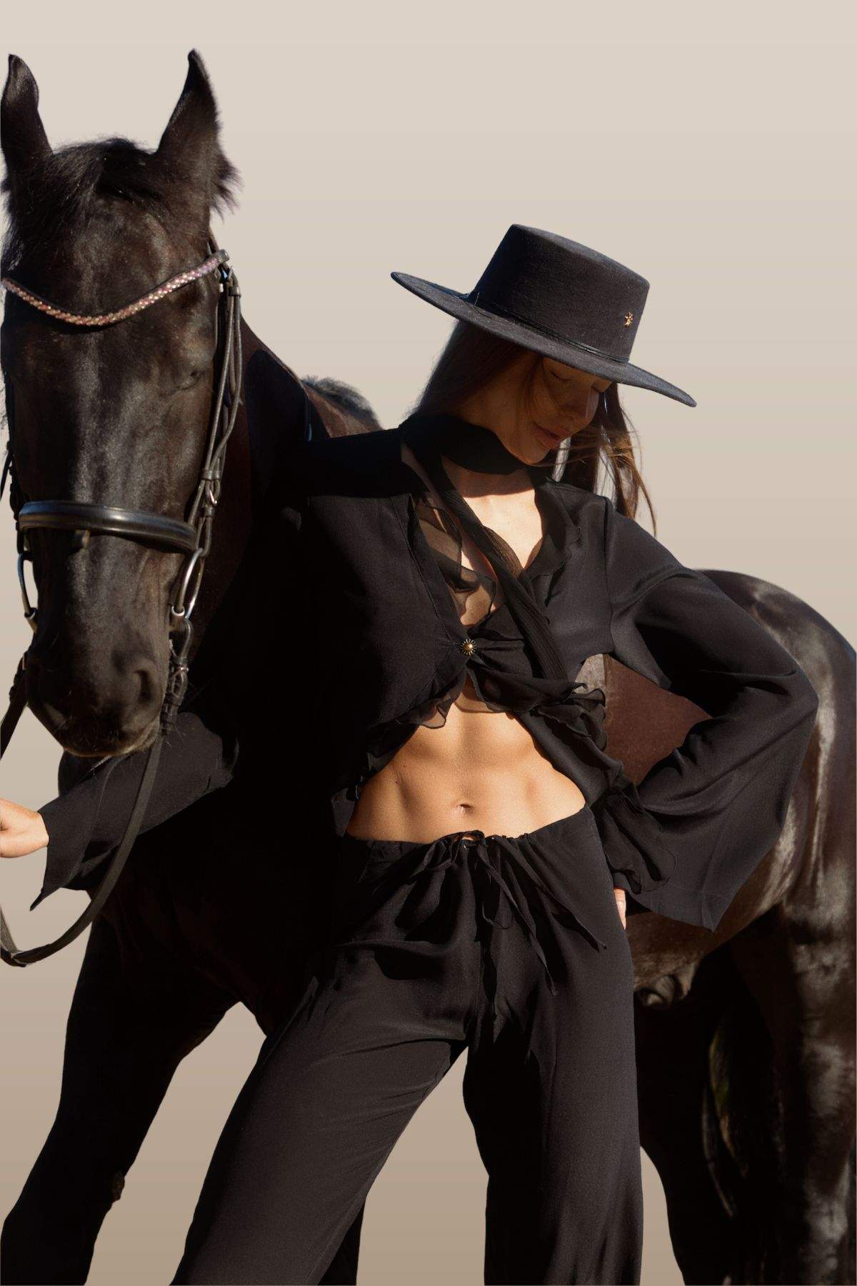 Woman in black outfit and hat standing next to a horse against a neutral background