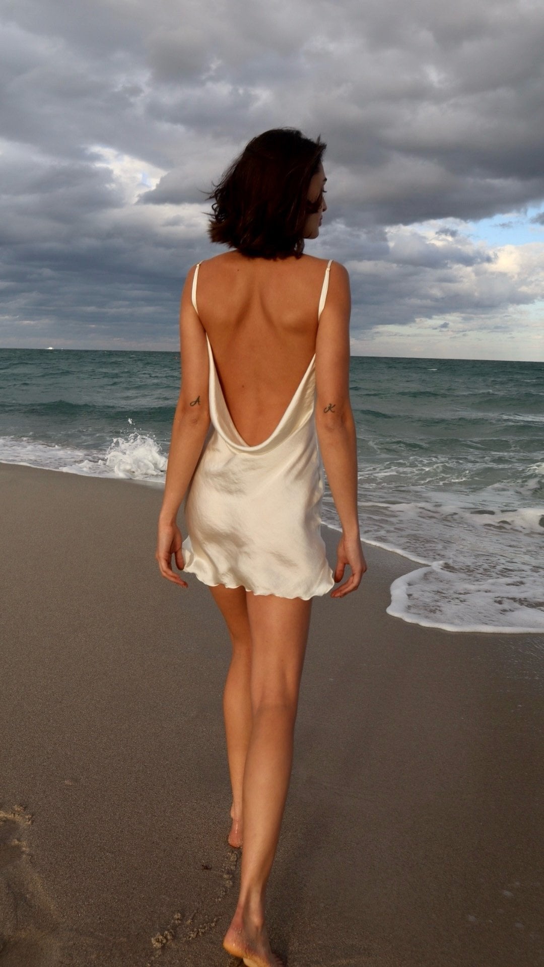 Woman in a white dress walking on a beach with a cloudy sky.