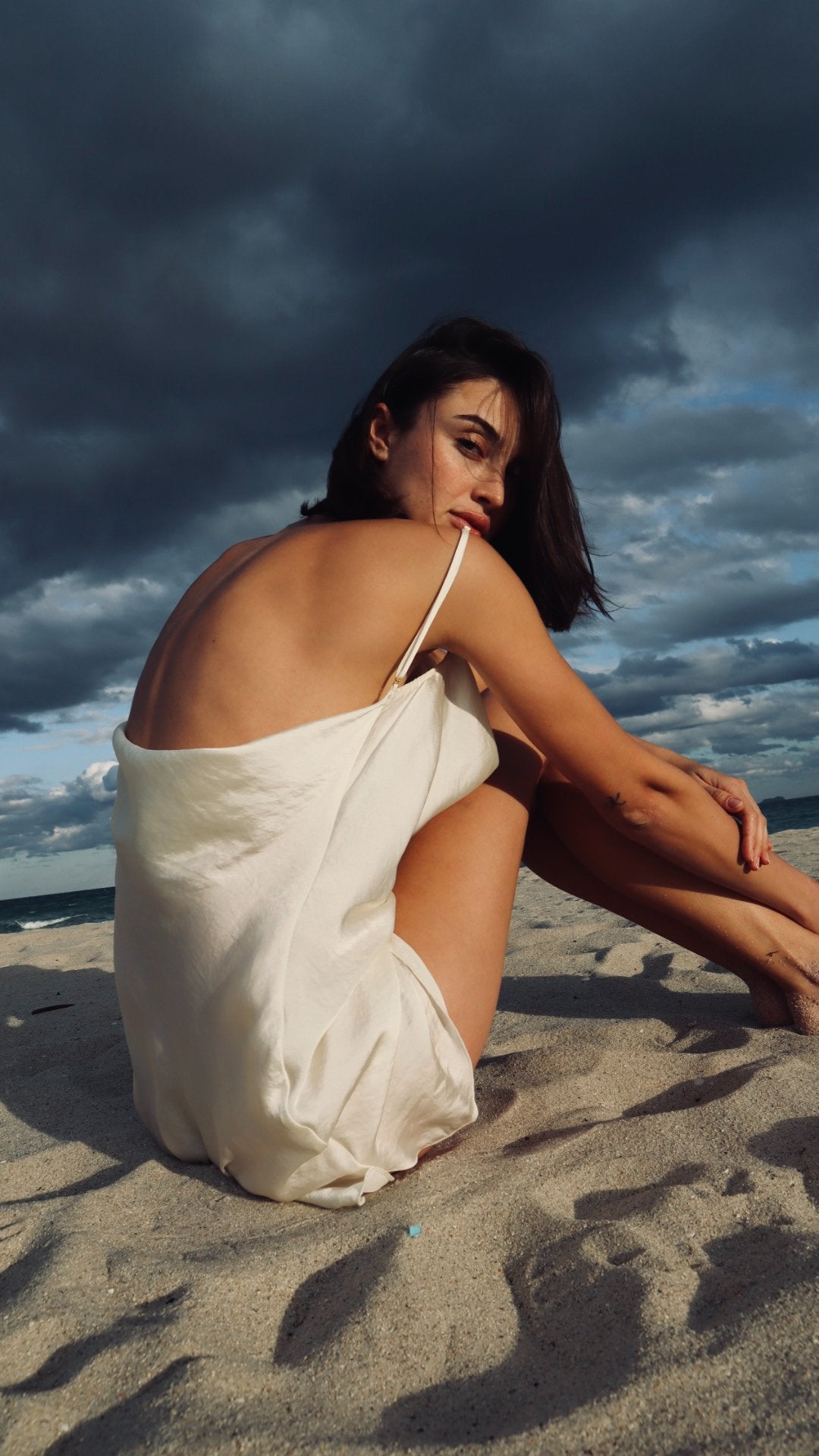 Woman in a white dress sitting on a beach with dark clouds in the sky