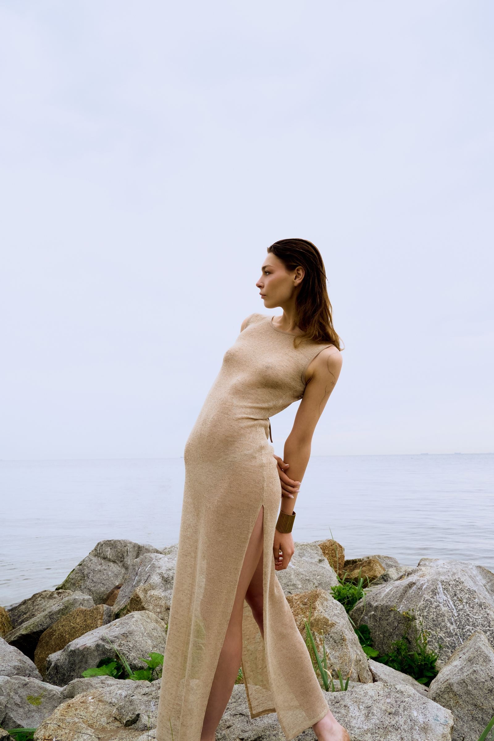 Woman in a beige dress standing on rocky terrain with a body of water in the background