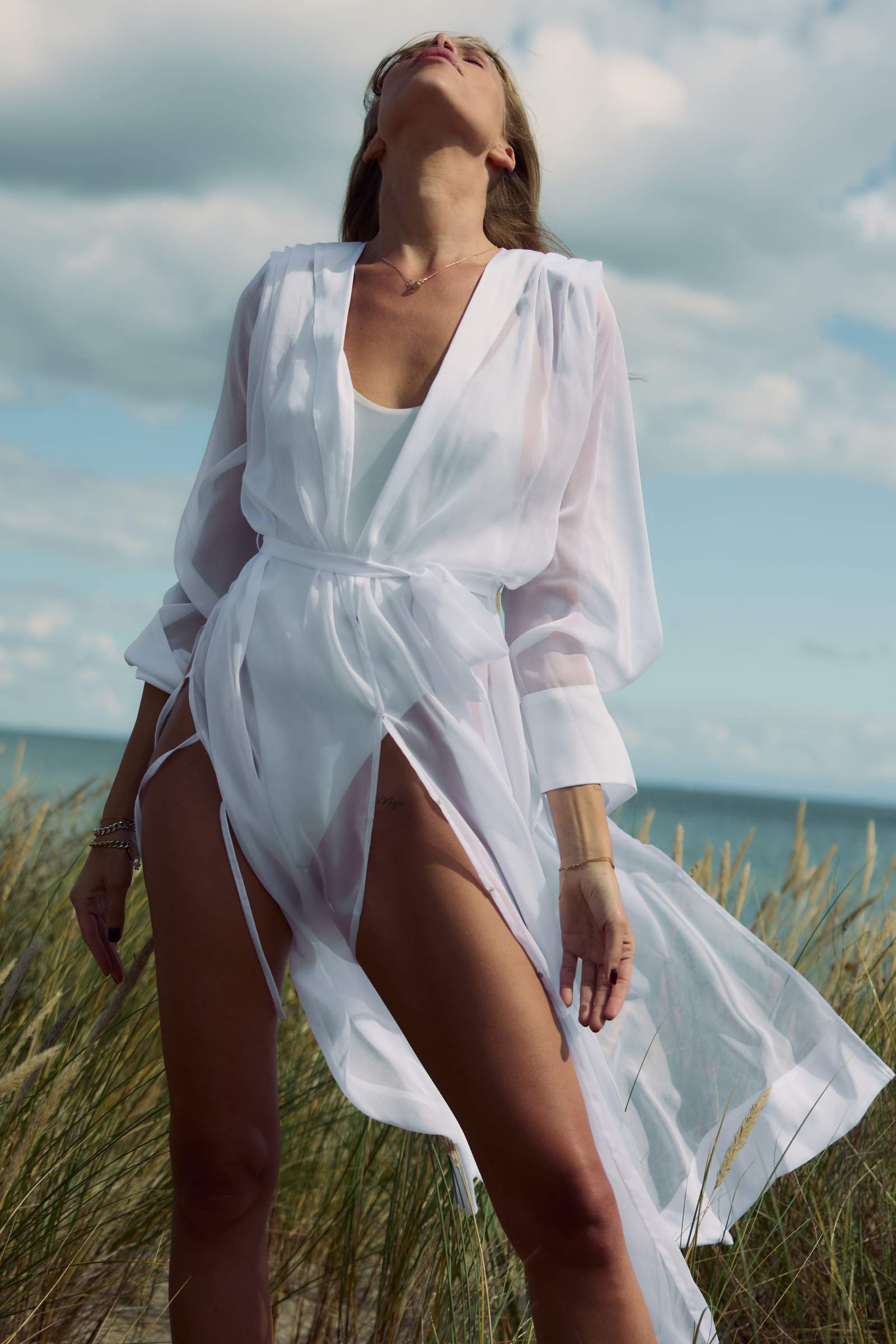 Woman in a white dress standing in tall grass with a cloudy sky background