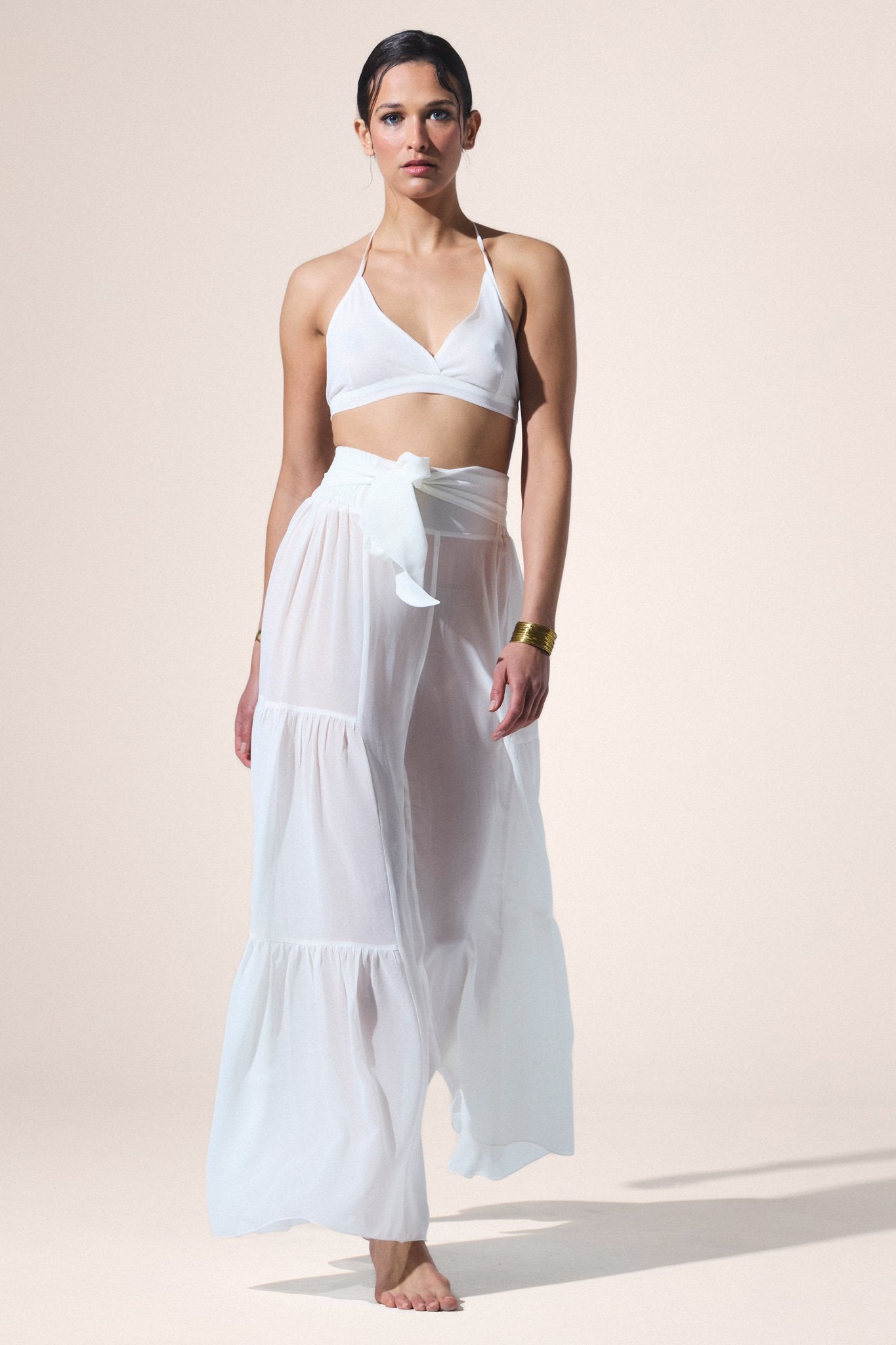 Woman wearing a white bikini top and flowing white skirt against a beige background
